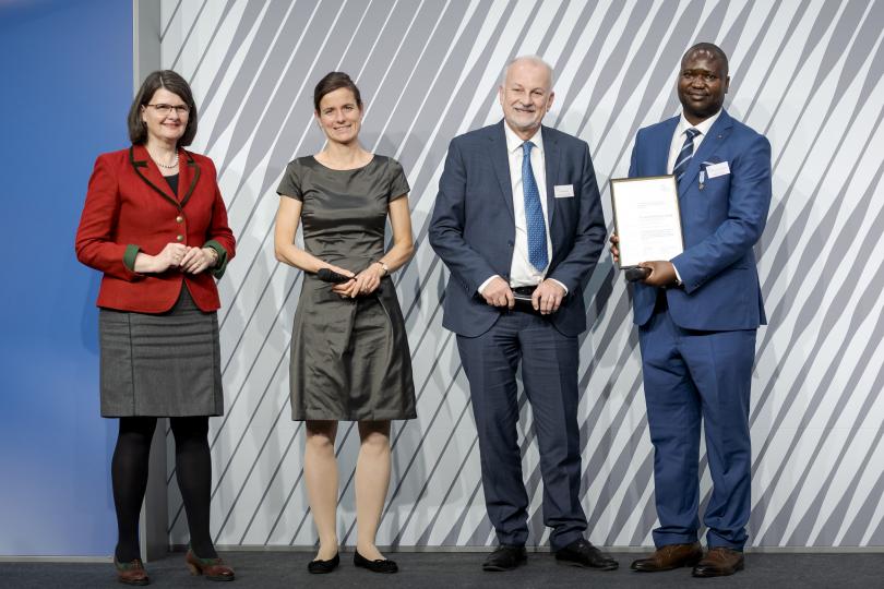 Dr. Jean-Paul Uvoyo Ulangi nimmt den Preis von Dr. Dieter Schenk entgegen – im Beisein von PD Dr. Carolin Kröner und Dr. Maria Flachsbarth.